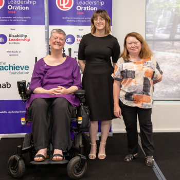 Three women posing together in front of Disability Leadership Institute Leadership Oration banners, one using a power wheelchair